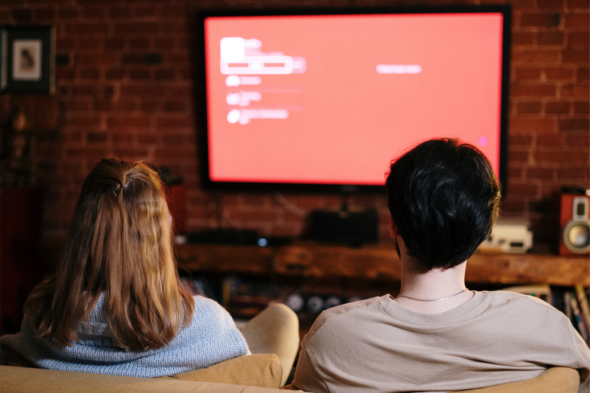 Na zdjęciu znajdują się dwie osoby. Kobieta i mężczyzna oglądający telewizję. Naprzeciwko nich jest ekran telewizora wyświetlający czerwone tło.